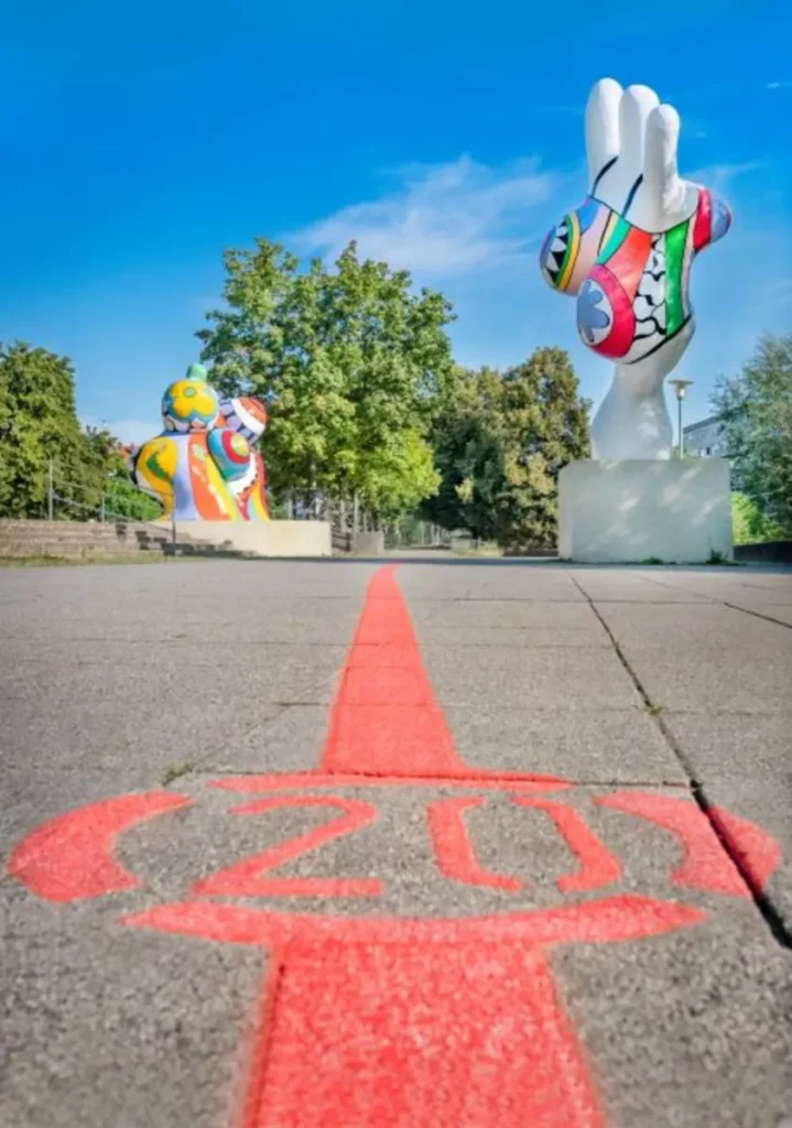 Die rote Linie des Roten Fadens mit der Stationsnummer 20 auf dem Gehweg, im Hintergrund die Nanas am Leibnizufer