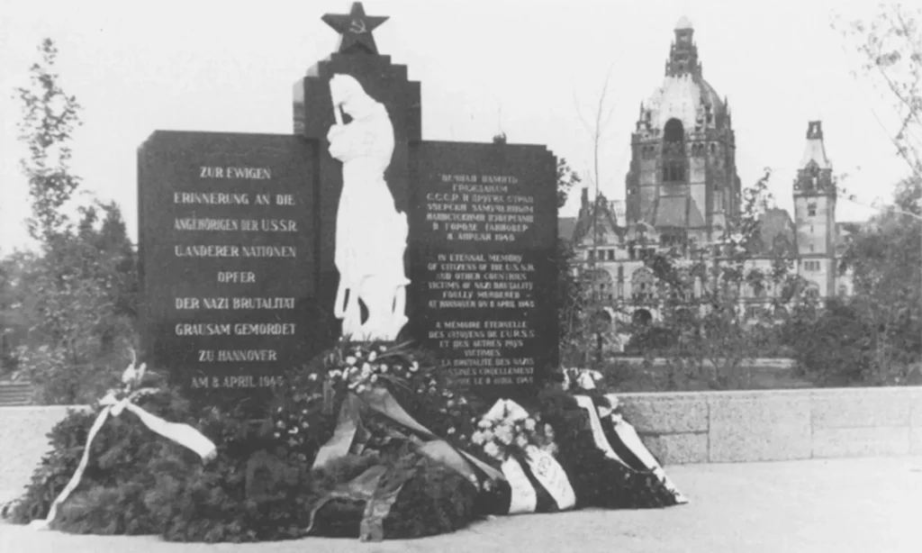 Historische Aufnahme des Gedenksteins für die ermordeten Opfer aus der Sowjetunion und anderen Nationen auf dem Ehrenfriedhof am Maschsee-Nordufer, davor Kränze, im Hintergrund das Neue Rathaus